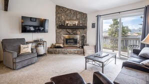 A living room with a stone fireplace, a flat-screen TV, and a balcony with a view of trees and mountains.