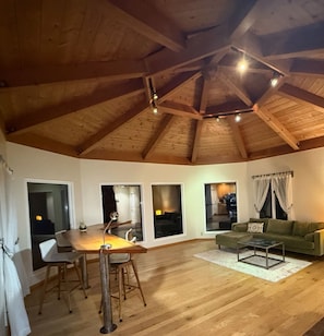 A spacious room with a wooden ceiling, a dining table with chairs, a sofa, and large windows.