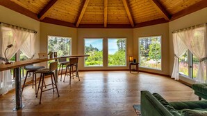 Une pièce spacieuse avec un haut plafond en bois, de grandes fenêtres donnant sur un paysage verdoyant, un coin repas avec une table et des chaises, et un canapé vert.