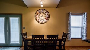 Une salle à manger avec une table et des chaises en bois, une horloge murale et une fenêtre ornée de rideaux à motifs.