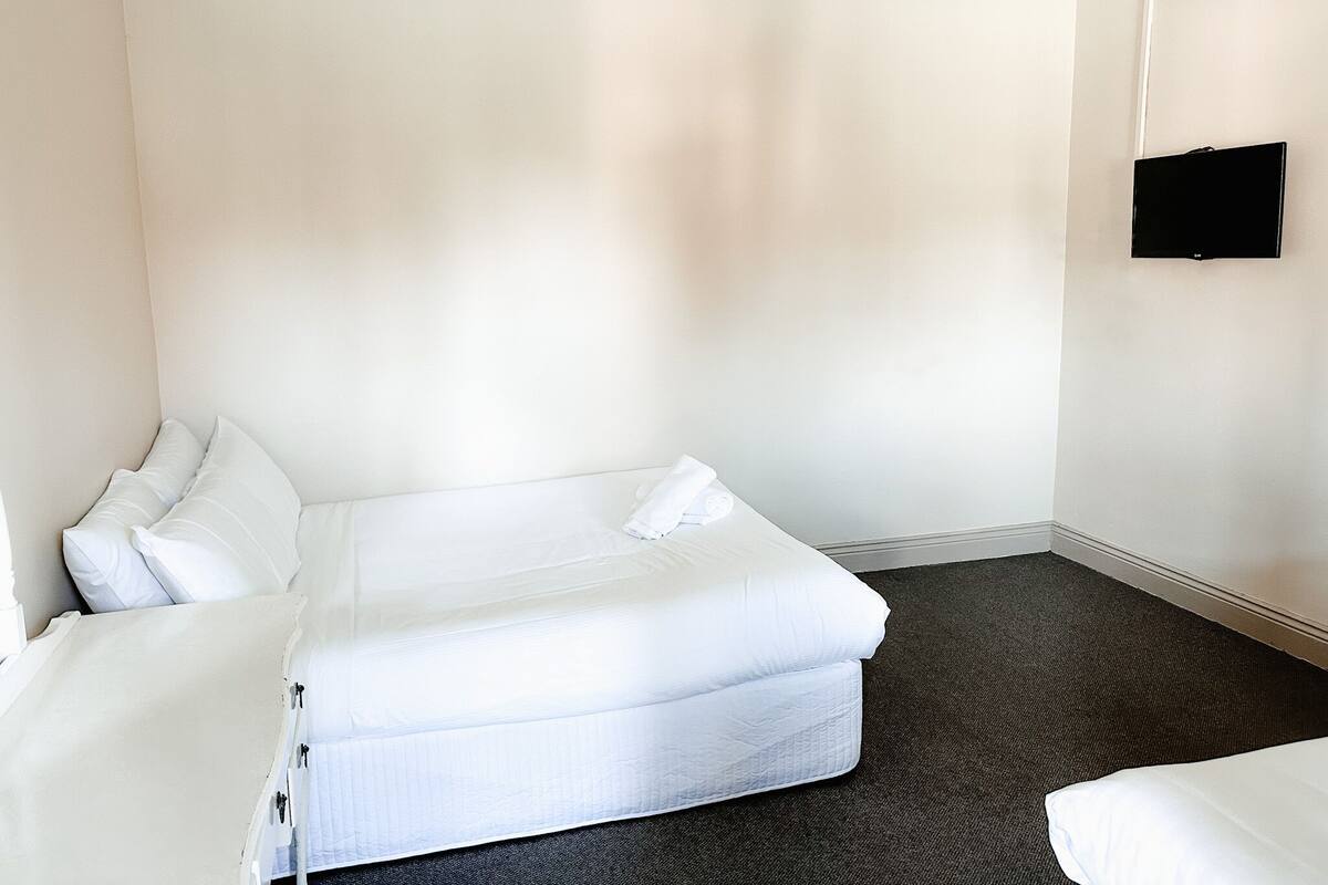 A minimalist hotel room with a single bed, a flat-screen TV mounted on the wall, and a dark carpet.