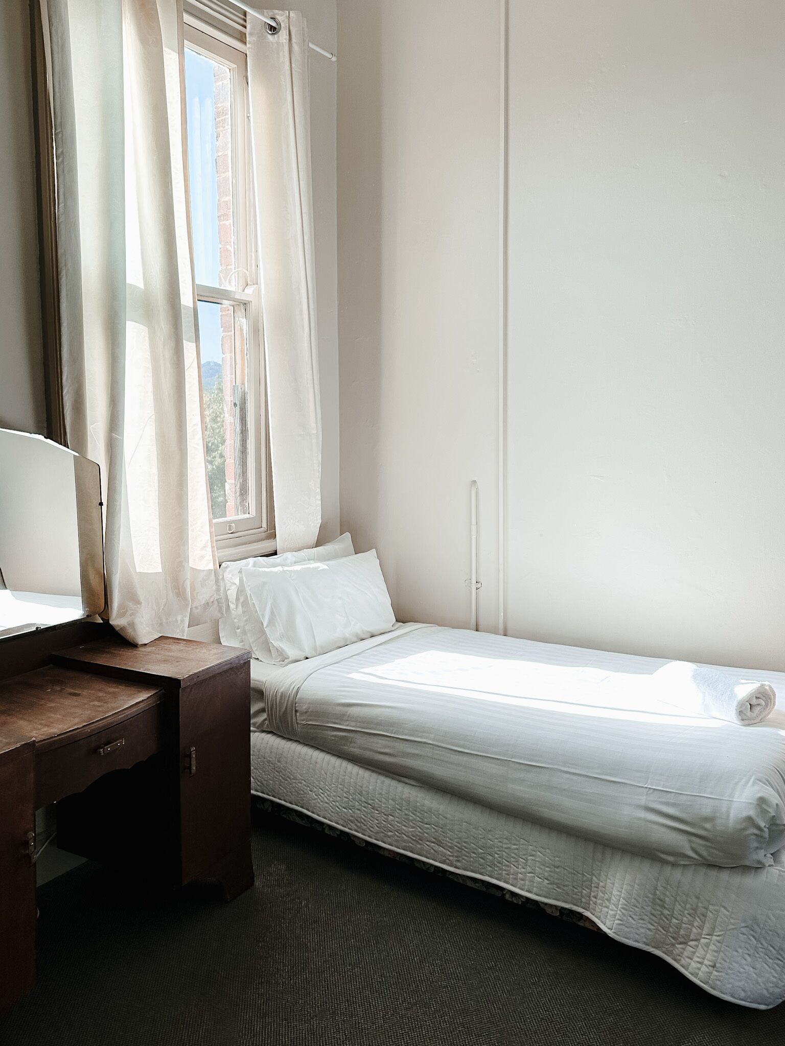A single bed with a white mattress and pillows, a wooden desk with a chair, and a window with curtains.