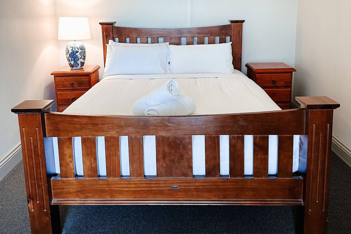A wooden bed with a white mattress and pillows, flanked by wooden nightstands with lamps. A towel is folded on the bed.