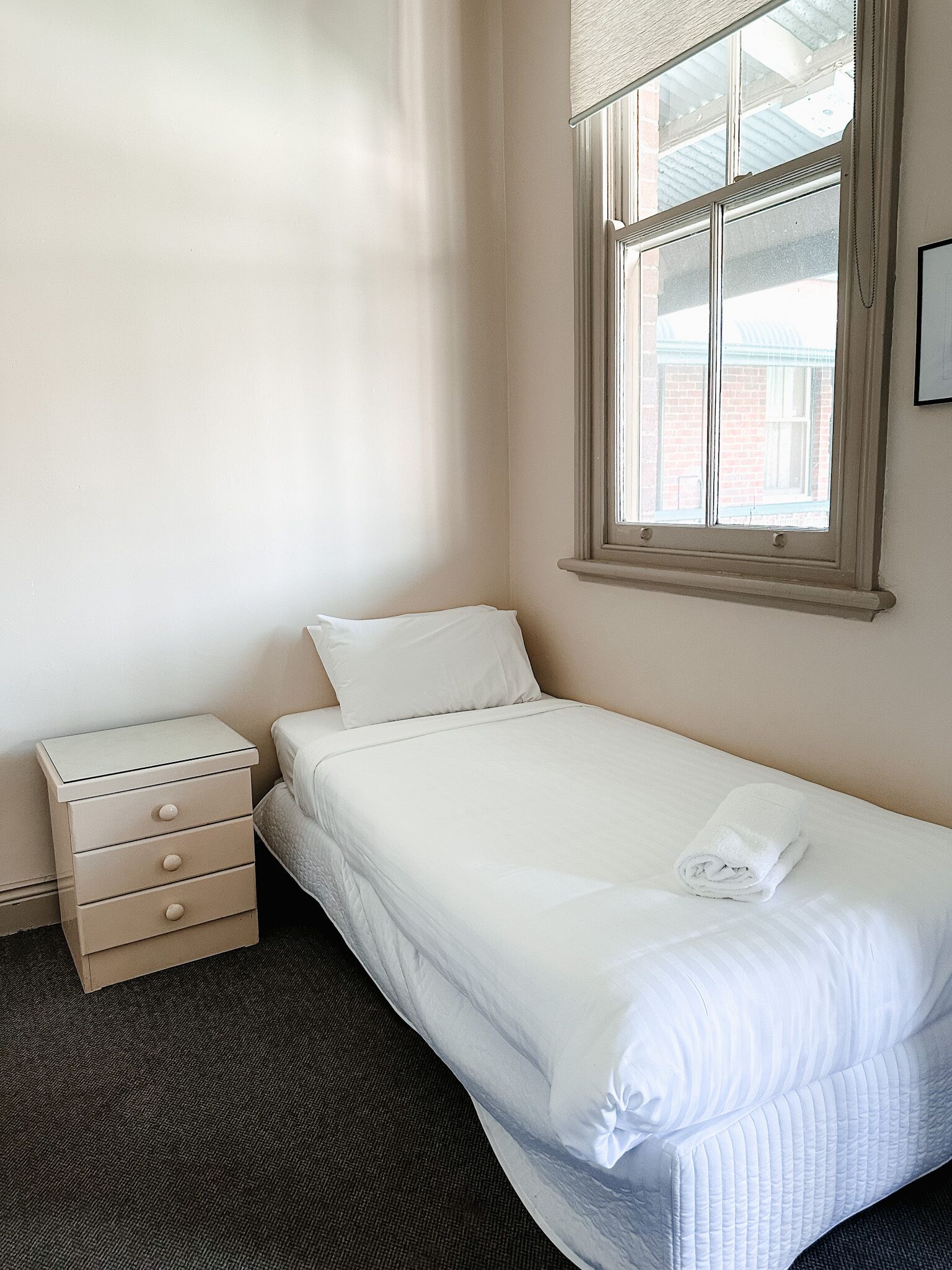 A small, neatly made bed with a white comforter and pillows, a wooden nightstand with drawers, and a window with a view of a brick building.