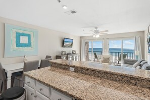 A kitchen with granite countertops, a dining area with a table and chairs, a ceiling fan, and a view of the ocean through large sliding doors.