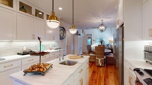 Una cocina moderna con armarios blancos, encimera de mármol y electrodomésticos de acero inoxidable. Hay un comedor con mobiliario de mimbre y vistas al salón.