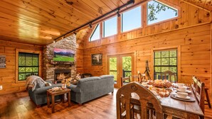 A cozy cabin interior with a stone fireplace, wooden walls, a sofa, a dining table set for four, and large windows.