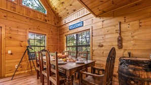A wooden cabin interior with a dining table set for four, a 'WELCOME' sign above a window, and a rustic barrel.