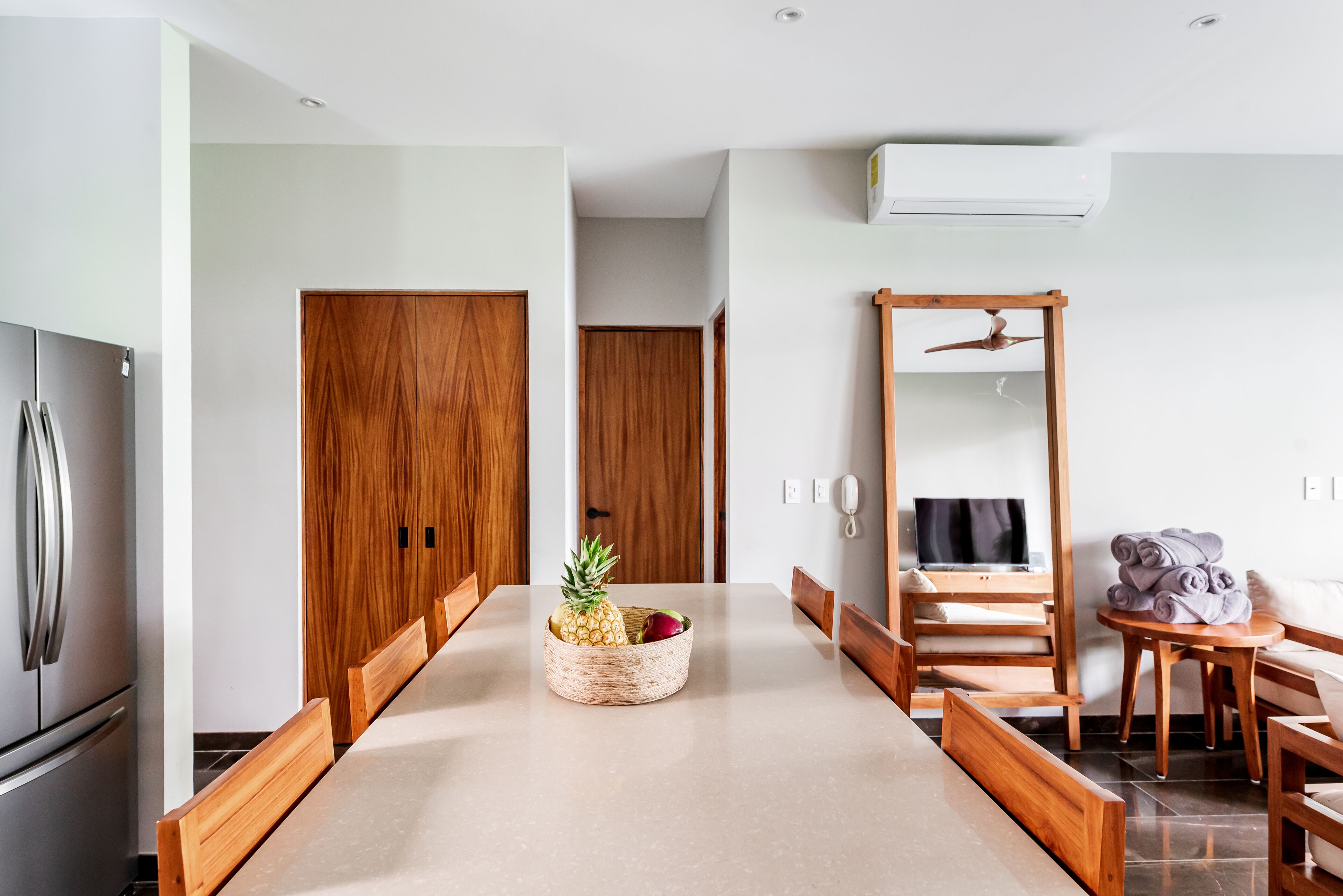 A modern kitchen with wooden cabinets, a stainless steel refrigerator, and a dining table with a woven bowl containing fruit.