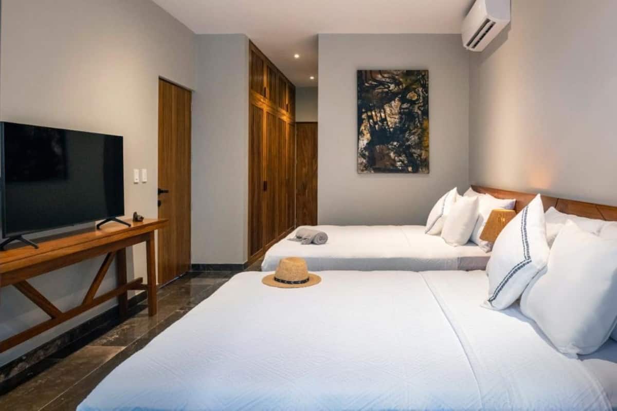 Una habitación de hotel con dos camas, un televisor sobre una mesa de madera, un cuadro en la pared y un equipo de aire acondicionado.