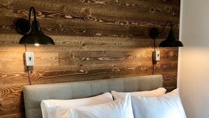Una cama con sábanas blancas, un cabecero gris y dos lámparas negras fijadas a la pared.