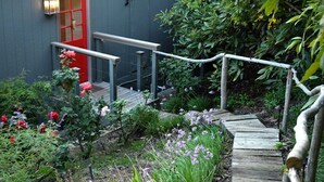 Una terraza de madera que conduce a una puerta roja, rodeada de diversas plantas y flores.