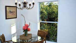 Un comedor con una mesa de vidrio, sillas de mimbre, un florero con flores, un cuadro enmarcado y una ventana con follaje en el exterior.