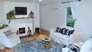 Una sala de estar con chimenea, sofás blancos, una mesa de centro de vidrio y un ventanal con cortinas.