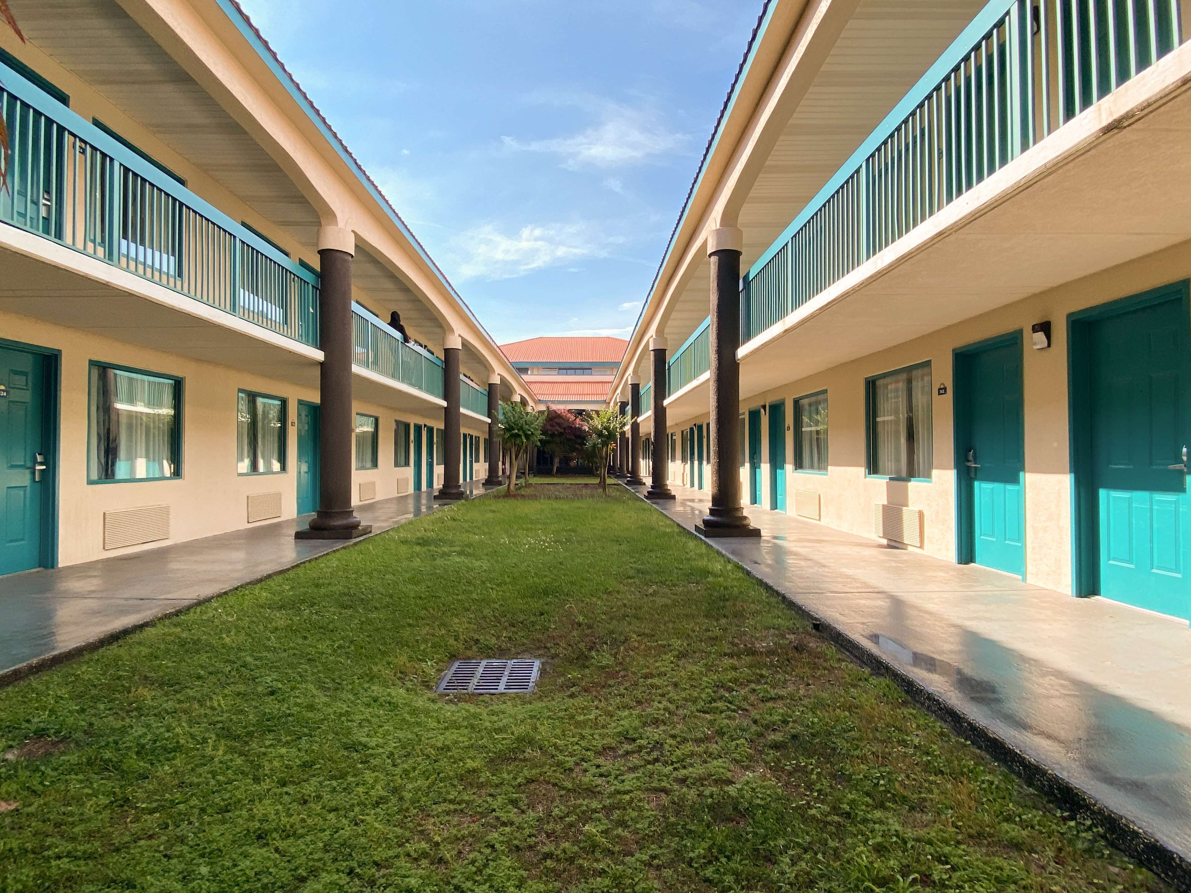 A corridor with a central lawn, flanked by buildings with teal doors and windows, leading to a red-roofed structure in the distance.