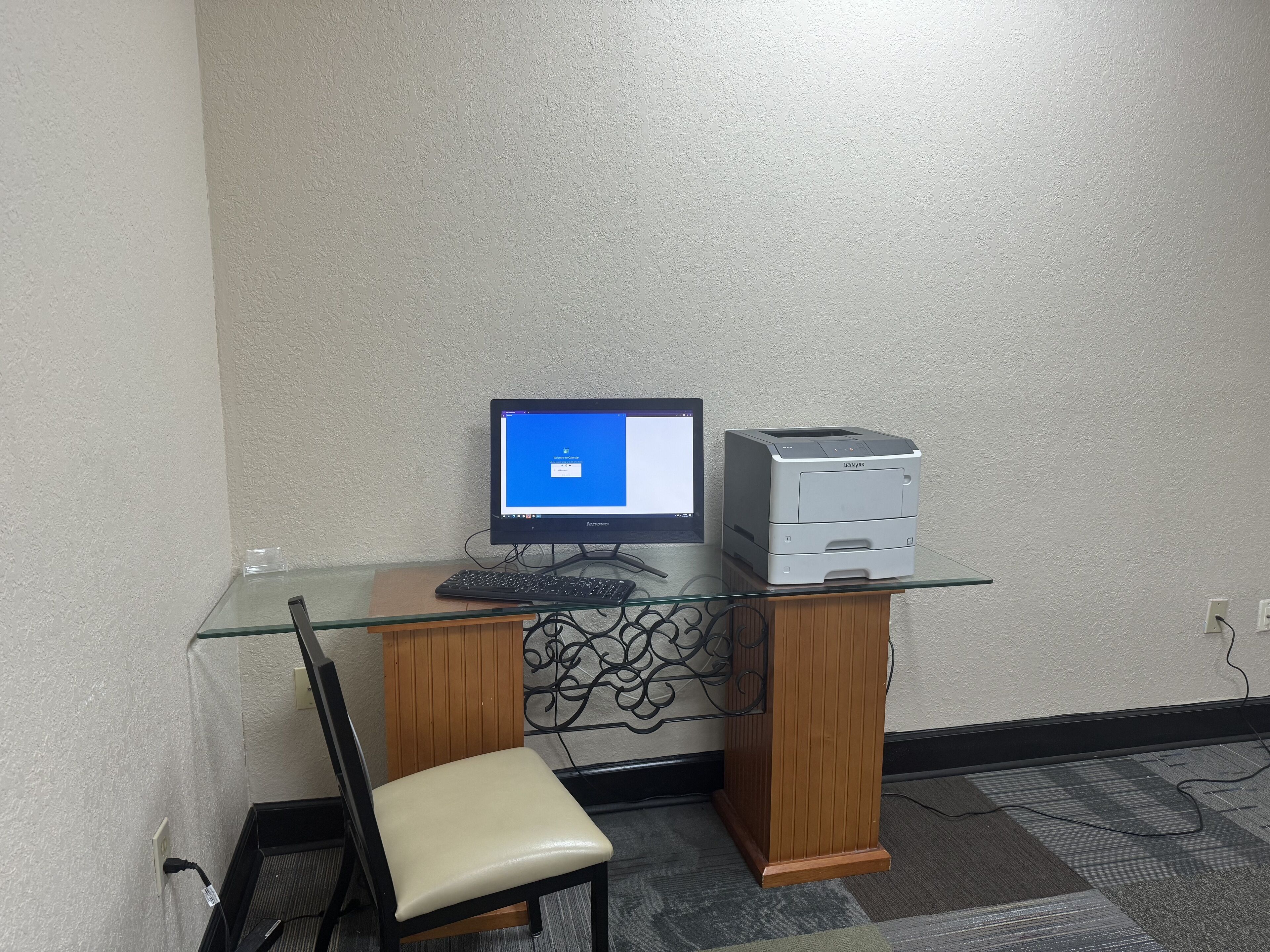 A corner office with a glass desk, a computer monitor, a printer, and a chair.