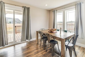 Et spiseområde med et træbord og fire stole, store vinduer med gardiner og udsigt til en baghave.