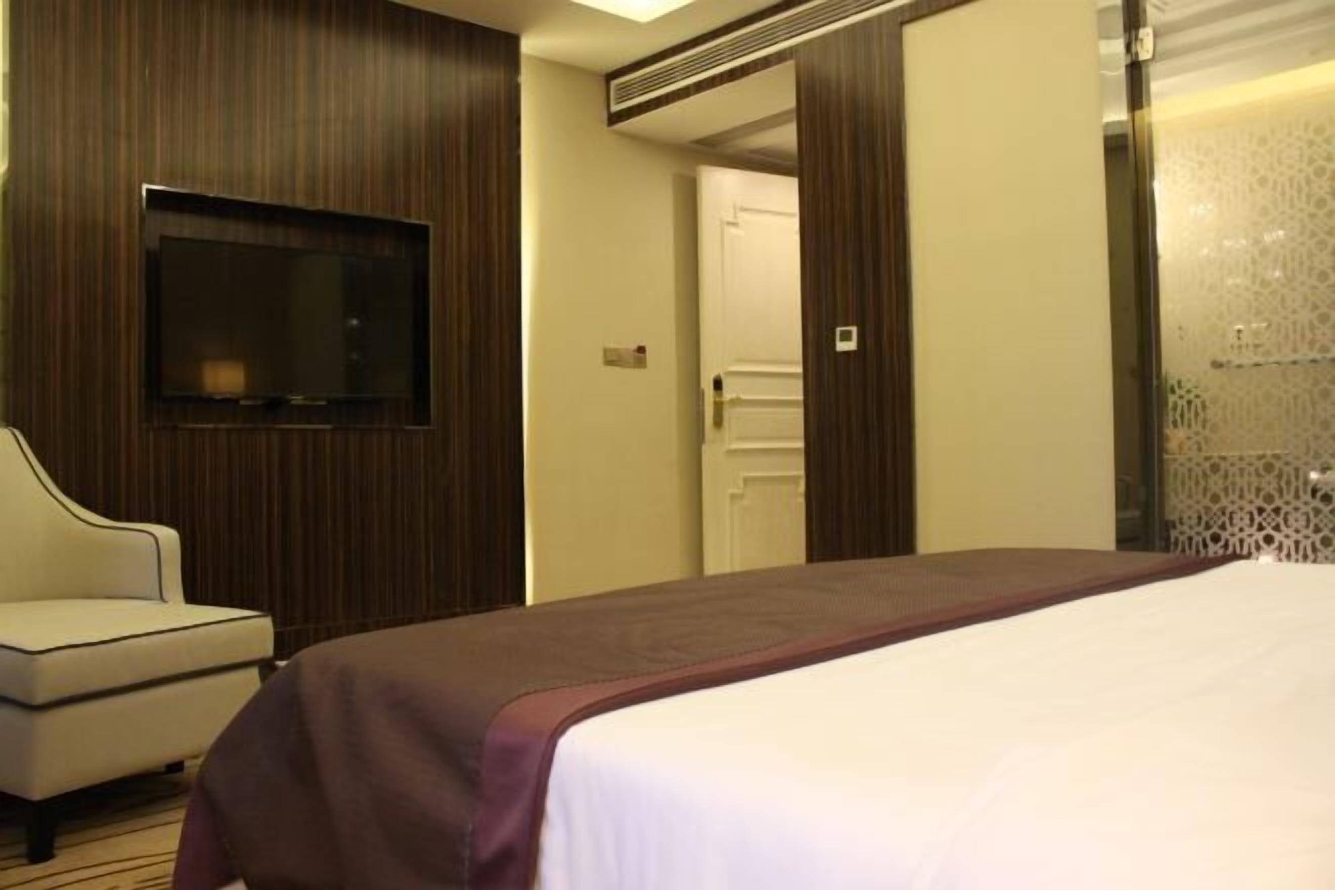 Ett hotellrum med en stor säng, en platt-TV som är monterad på väggen, en sittgrupp med en stol och en dörr som leder till ett annat rum.
