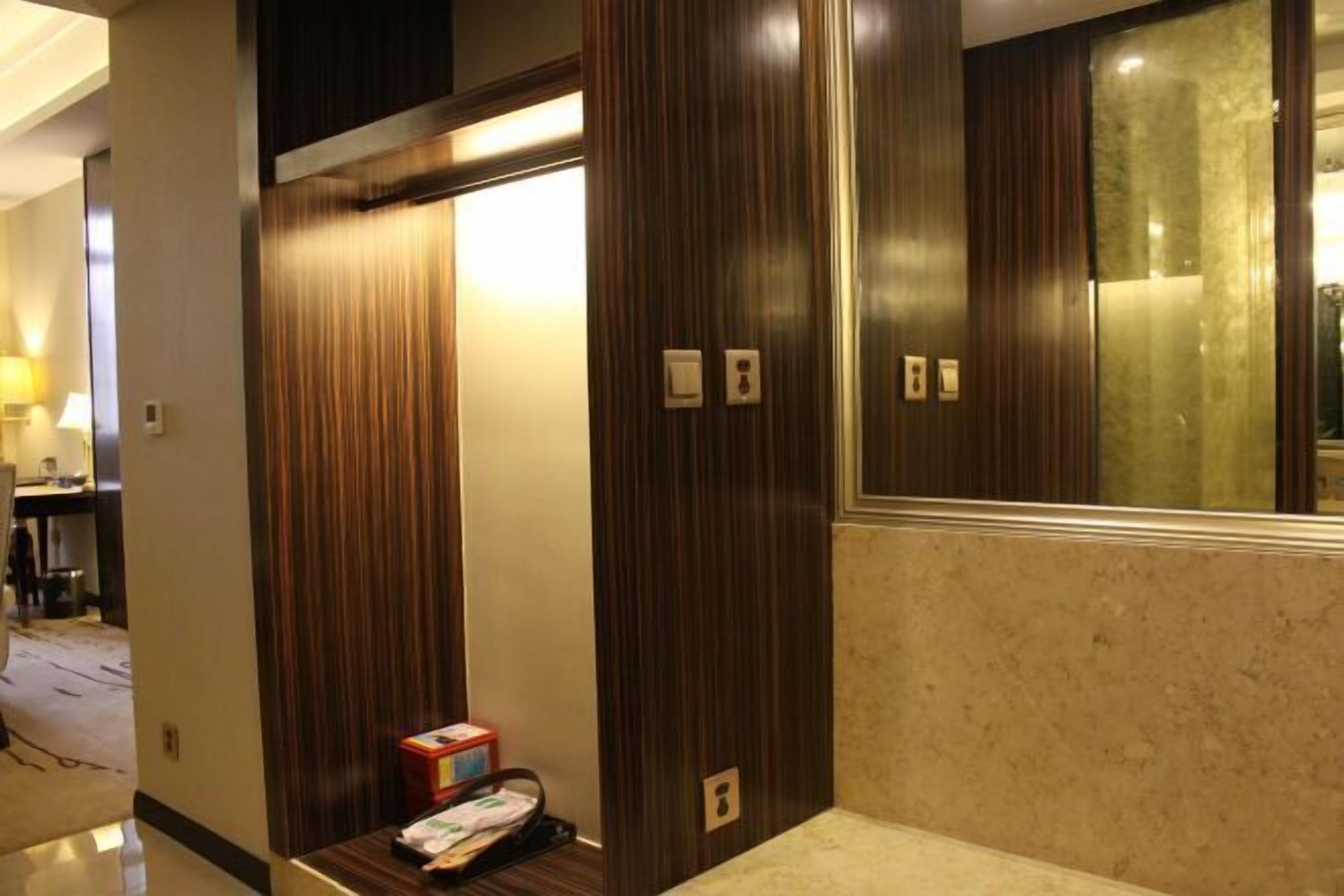 A hotel room with a wooden wardrobe, a small table with a box and papers, and a mirror with a wooden frame.