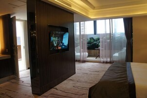 Habitación de hotel con una cama grande, un televisor instalado en un panel de madera, un balcón con vistas a zonas verdes y suelo alfombrado.