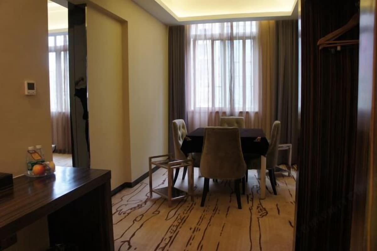 A hotel room with a dining area featuring a table and chairs, a wooden floor, and a view through sheer curtains.