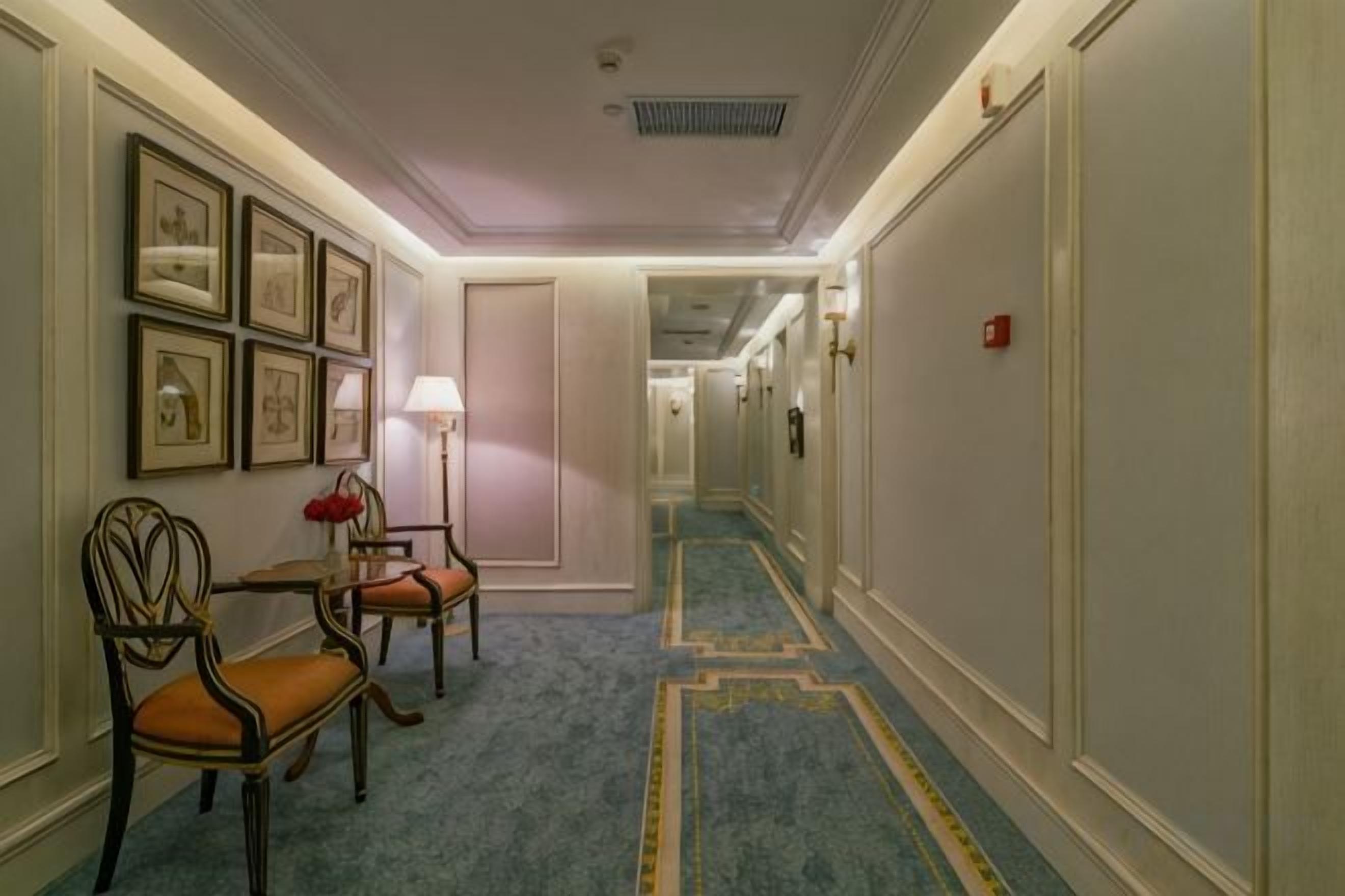 A hotel hallway with a carpeted floor, framed pictures on the wall, a lamp, and a fire alarm.