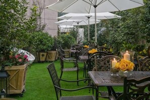 Um pátio externo com mesas e cadeiras, cercado por vegetação e guarda-sóis.