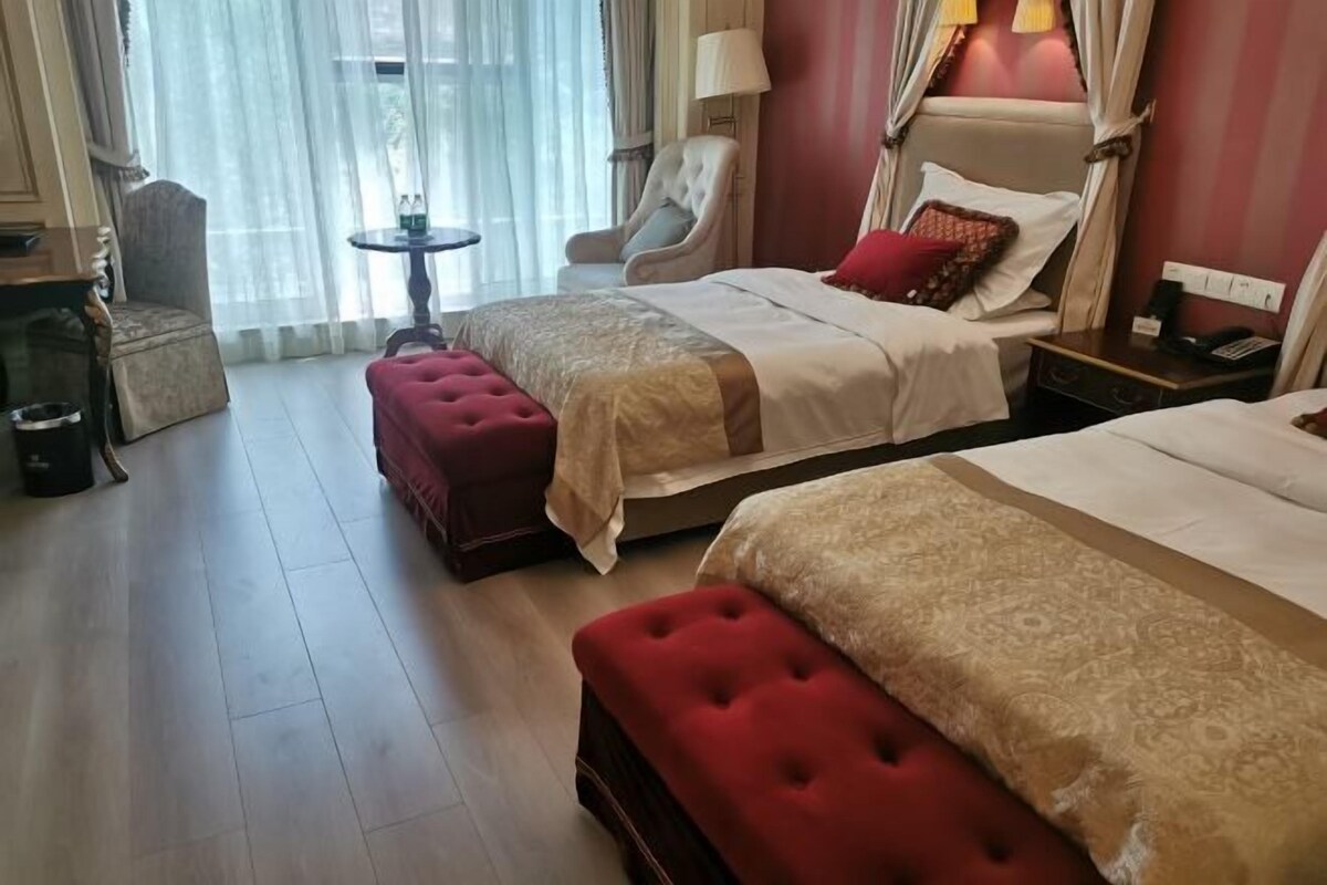 A hotel room with two beds, a red upholstered bench, a small round table, and a chair.