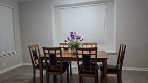 Ruokailuhuoneessa on puinen pöytä ja tuolit, maljakko, jossa on violetteja kukkia, sekä ikkuna, jossa on sälekaihtimet.