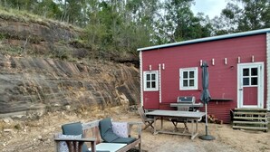 A rustic outdoor seating area with a sofa and table, a red building with windows and a door, and a rocky hillside in the background.