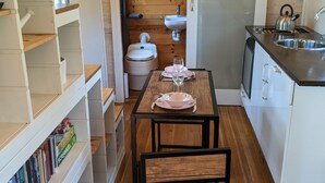 A compact kitchen with a wooden table, two chairs, a sink, and a toilet. Shelves with books and a potted plant are visible.