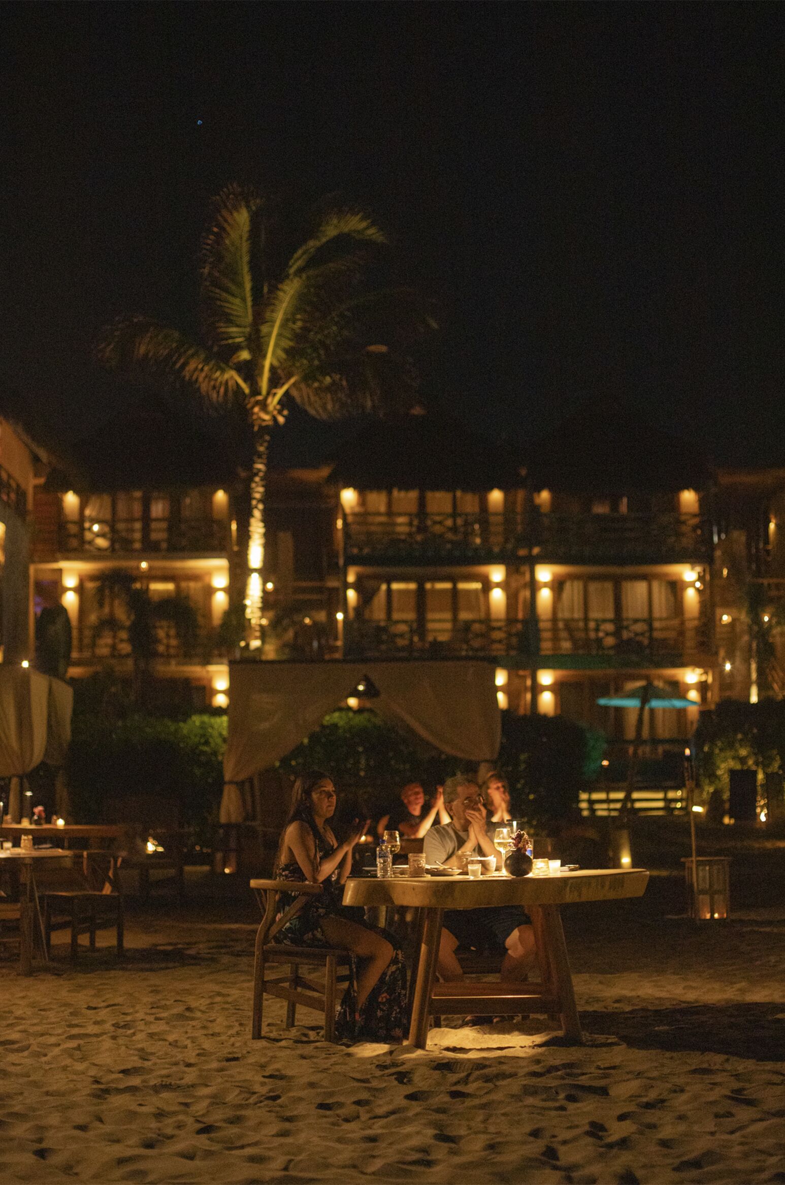 A beachfront dining area with tables and chairs, palm trees, and a building with multiple windows and lights in the background.