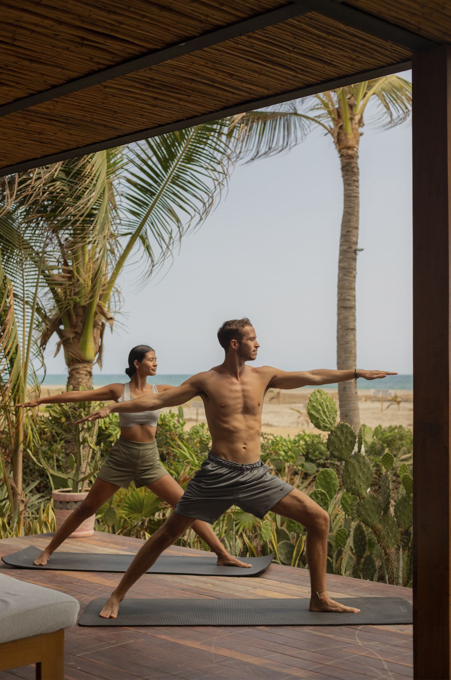 Dos personas practicando yoga sobre esterillas al aire libre, con una playa y palmeras al fondo.