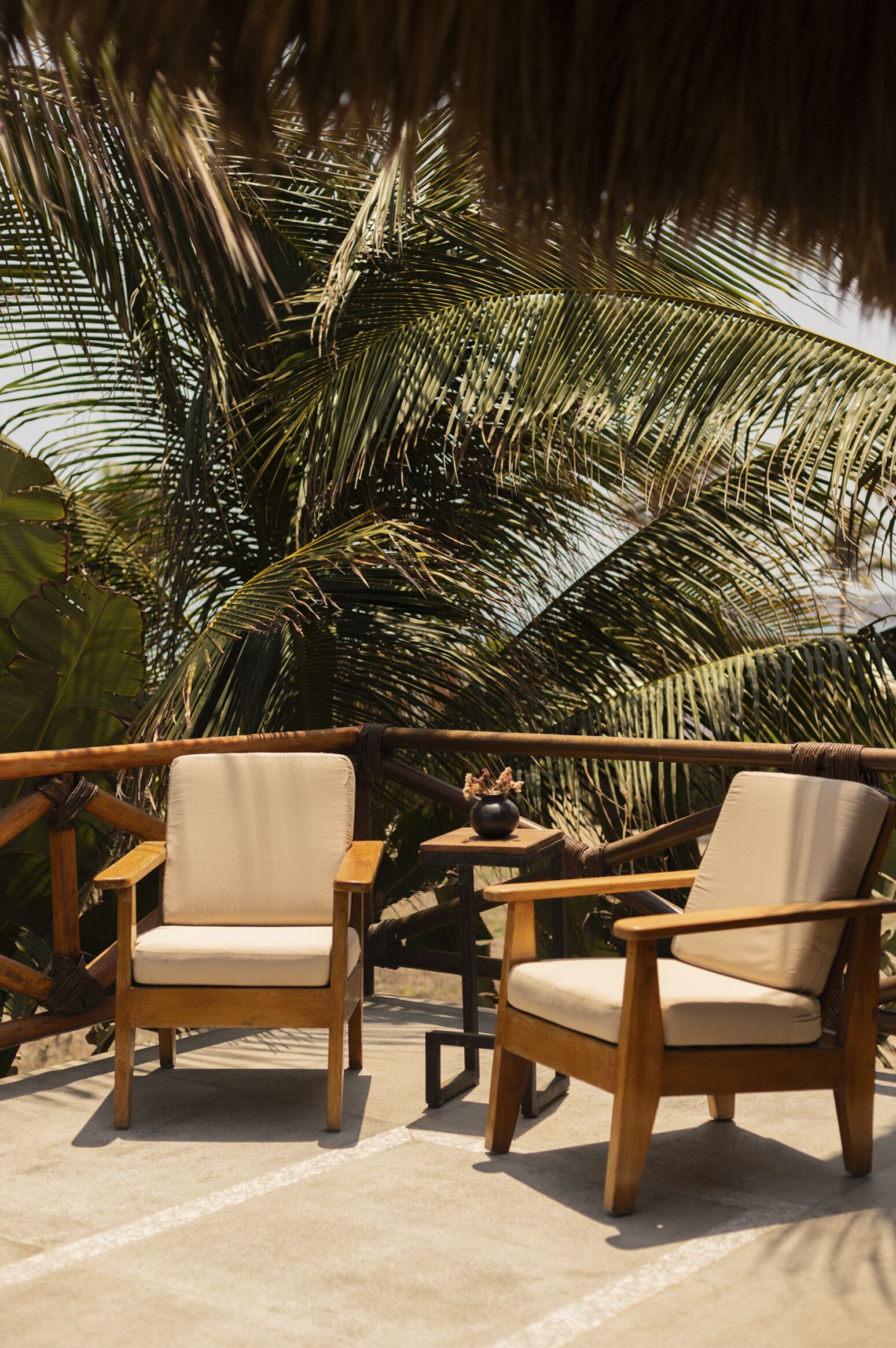 A wooden deck with two beige cushioned chairs and a small table, surrounded by lush palm trees.