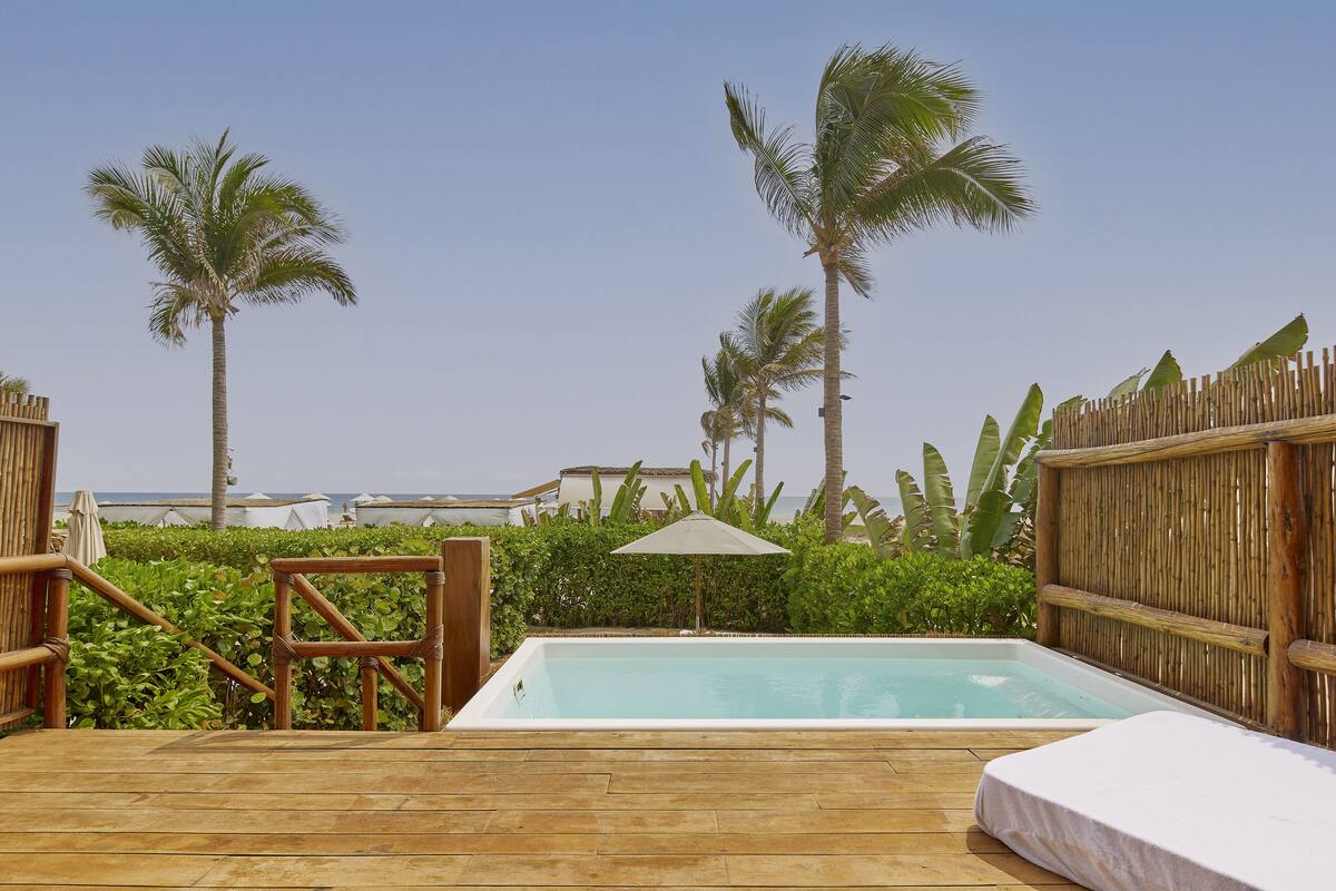 Une terrasse en bois dotée d’un jacuzzi, entourée d’une végétation luxuriante et de palmiers, avec vue sur une plage.
