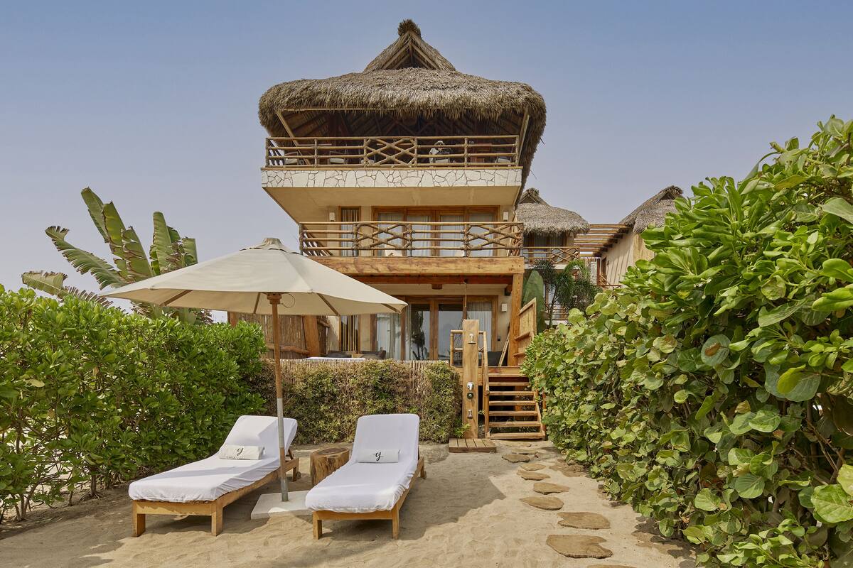 Une villa située en bord de mer, dotée d’un toit de chaume, d’un balcon avec des balustrades en bois et d’un chemin de sable menant à un espace de détente avec deux chaises longues et un parasol.