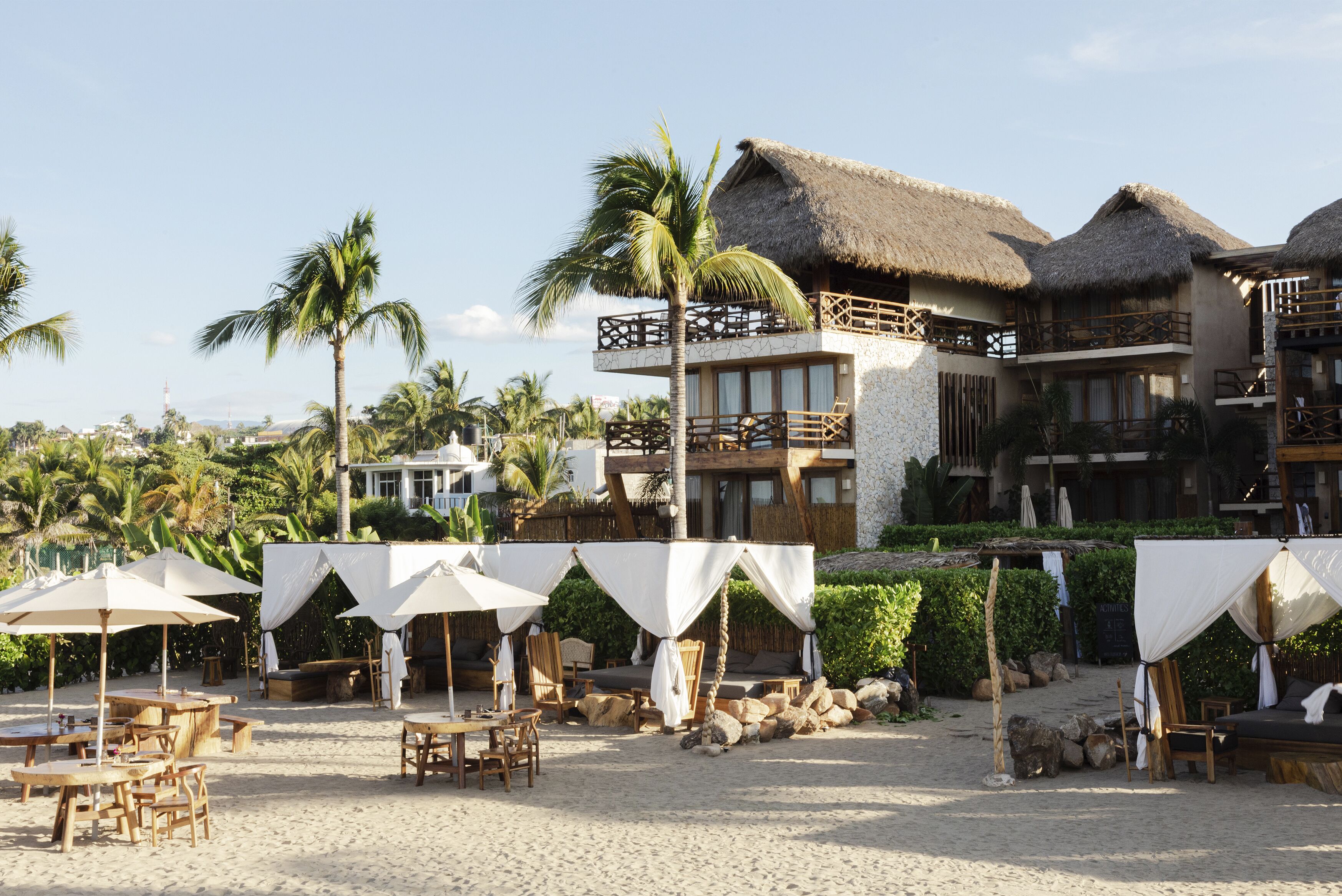 Un resort frente a la playa con edificios de techo de paja, mobiliario exterior y palmeras.