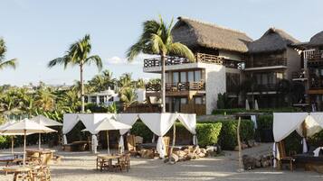 Un resort frente a la playa con edificios de techo de paja, mobiliario exterior y palmeras.