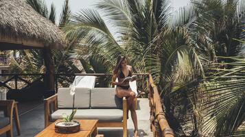 A woman sitting on a sofa in a tropical setting with palm trees and a thatched roof structure.