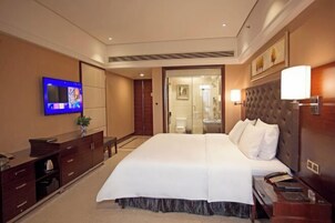 A hotel room with a large bed, a flat-screen TV mounted on the wall, a desk with a chair, and a bathroom visible through an open door.