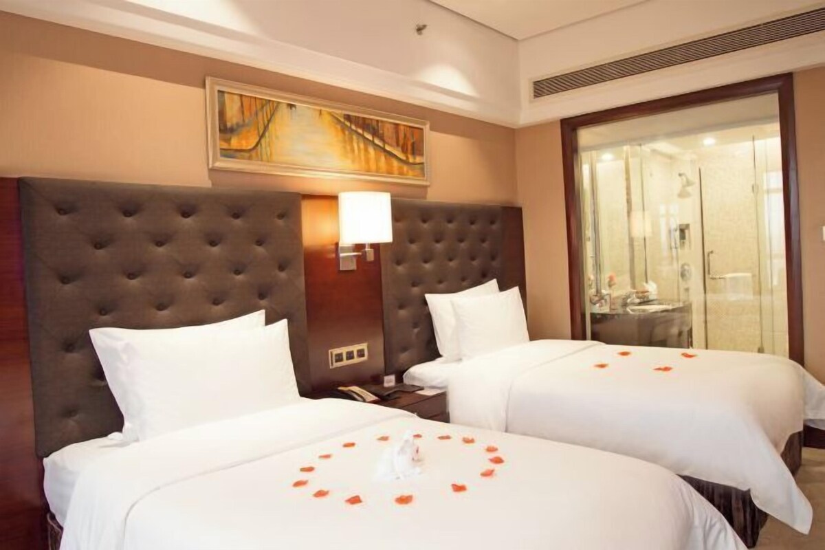 A hotel room with two beds, a large headboard, a painting on the wall, and a bathroom visible through a glass door.
