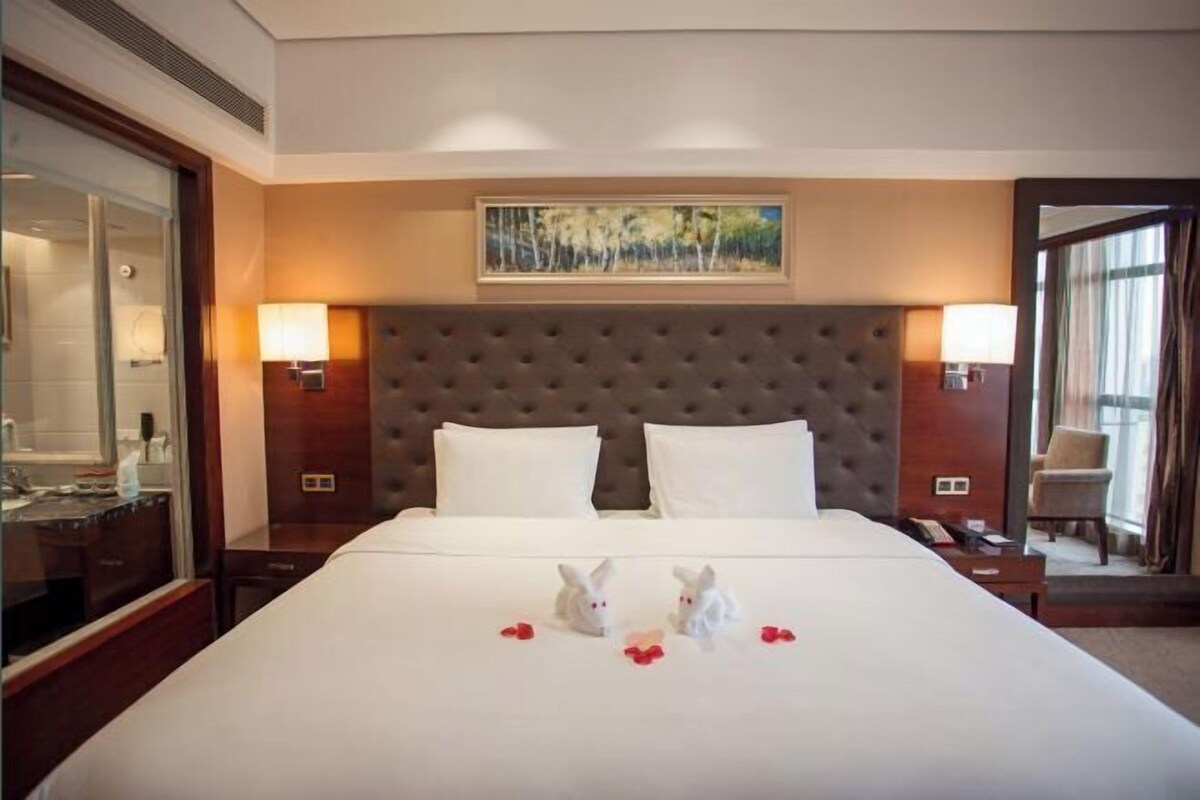A hotel room with a large bed, two bedside lamps, a headboard, and a painting above the bed.