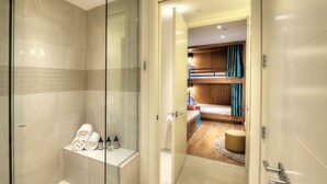 Ein modernes Badezimmer mit Duschkabine aus Glas, einem Waschtisch mit Pflegeprodukten und Blick in ein Schlafzimmer mit Etagenbett und Holz-Kopfteil.