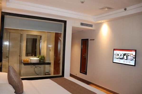 A hotel room with a large sliding glass door leading to a bathroom, a flat-screen TV mounted on the wall, and a bed with a pillow.