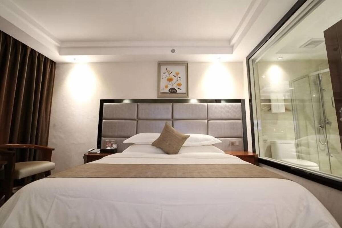Ein Hotelzimmer mit einem großen Bett, einem Nachttisch, einem Sessel, einer Duschkabine aus Glas und einem gerahmten Bild an der Wand.