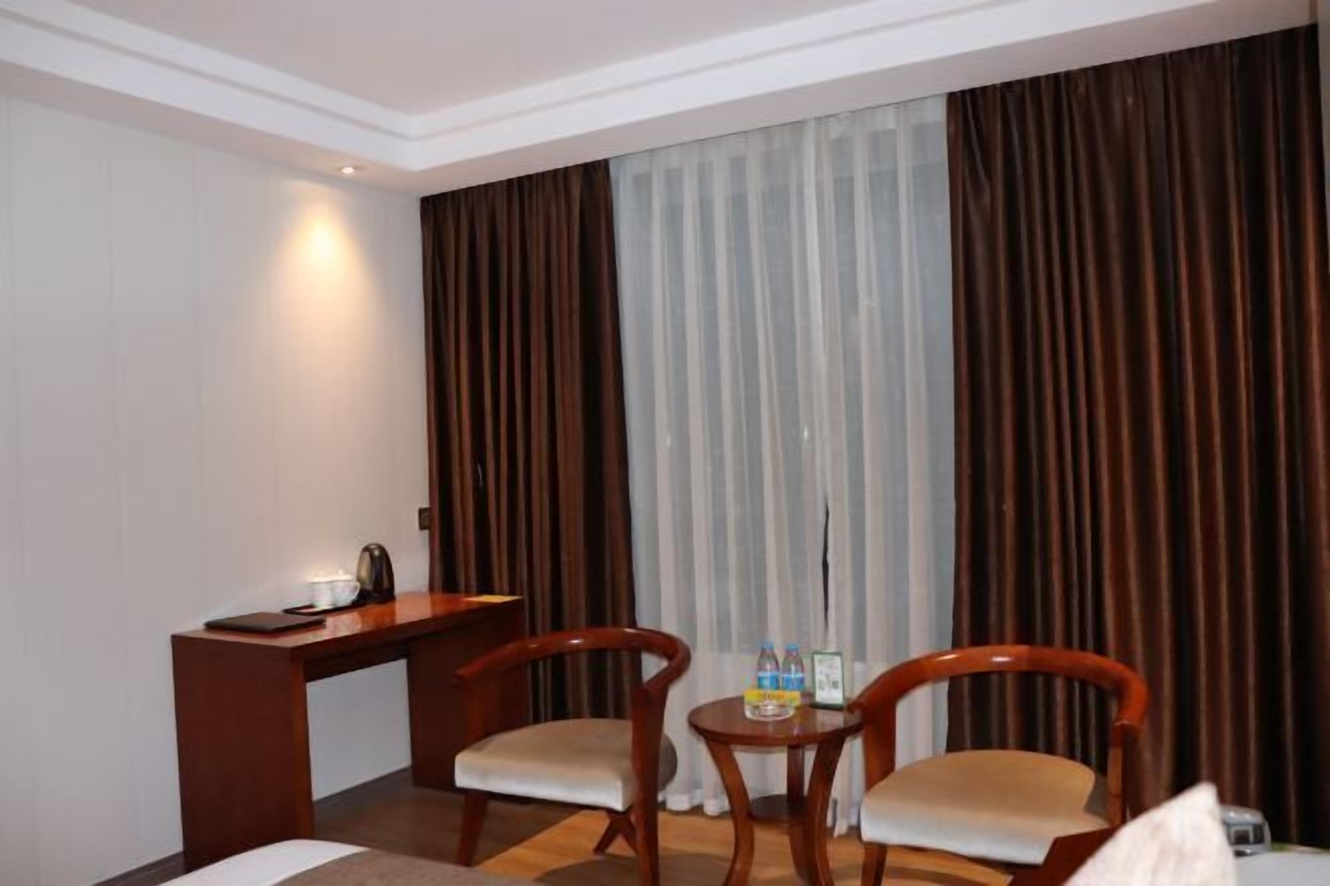Habitación de hotel con escritorio de madera, dos sillas, una mesita con botellas de agua y un ventanal con cortinas.
