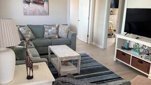 A living room with a grey sofa, a white coffee table, a wicker chair, a ceiling fan, and a painting of boats on the wall. - Oak Island Beach House -- Sand, Sun, and Fun Just Steps Away!! (Oak Island)