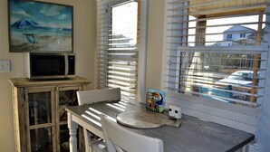 A dining area with a wooden table, white chairs, a microwave on a cabinet, and a painting of a beach scene. - Oak Island Beach House -- Sand, Sun, and Fun Just Steps Away!! (Oak Island)
