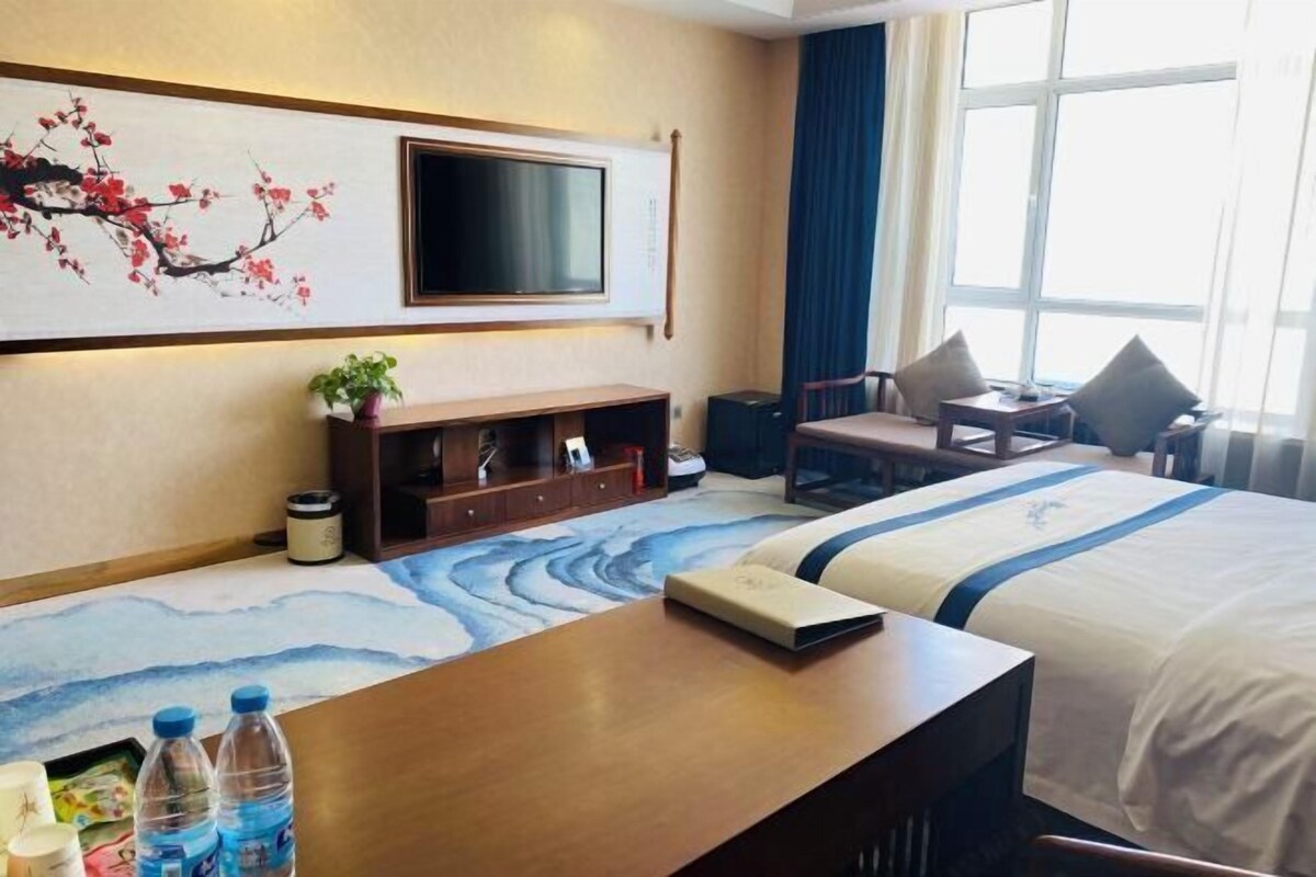 A hotel room with a bed, a TV mounted on the wall, a desk with a chair, and a window with curtains.