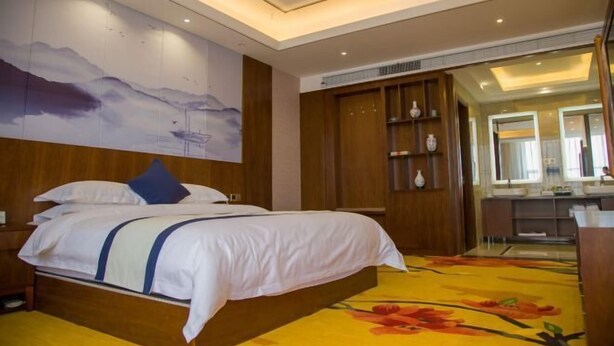 Una habitación de hotel con una cama, un cuadro grande en la pared, un armario de madera y un baño visible a través de una puerta abierta.